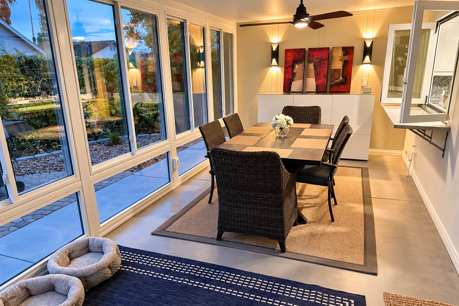 A custom sunroom dining area with a long table and wicker chairs beside broad windows and an open glass door.