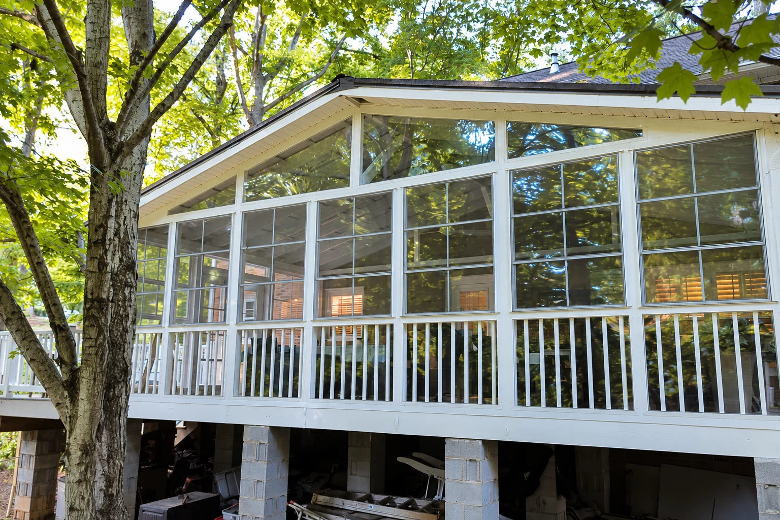 Raised deck enclosed as a white-framed sunroom, with large windows beneath a peaked roof among trees.