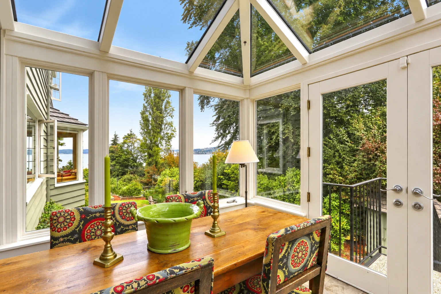A bright solarium with a glass roof and white frames contains a wooden dining table and patterned chairs, with trees and water visible outside.