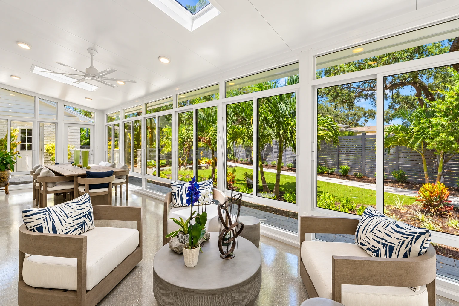 A sunroom with white-framed windows and skylights has lounge seating and a dining table overlooking a leafy garden.