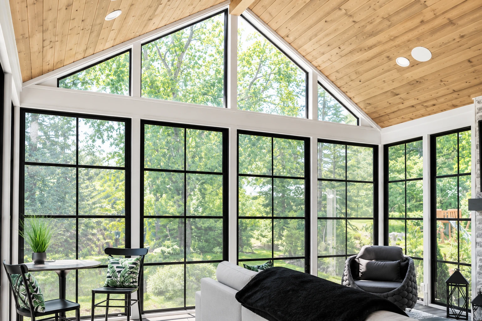 A bright sunroom with a peaked wood ceiling, tall black-framed windows, and a sofa and chairs overlooking trees.