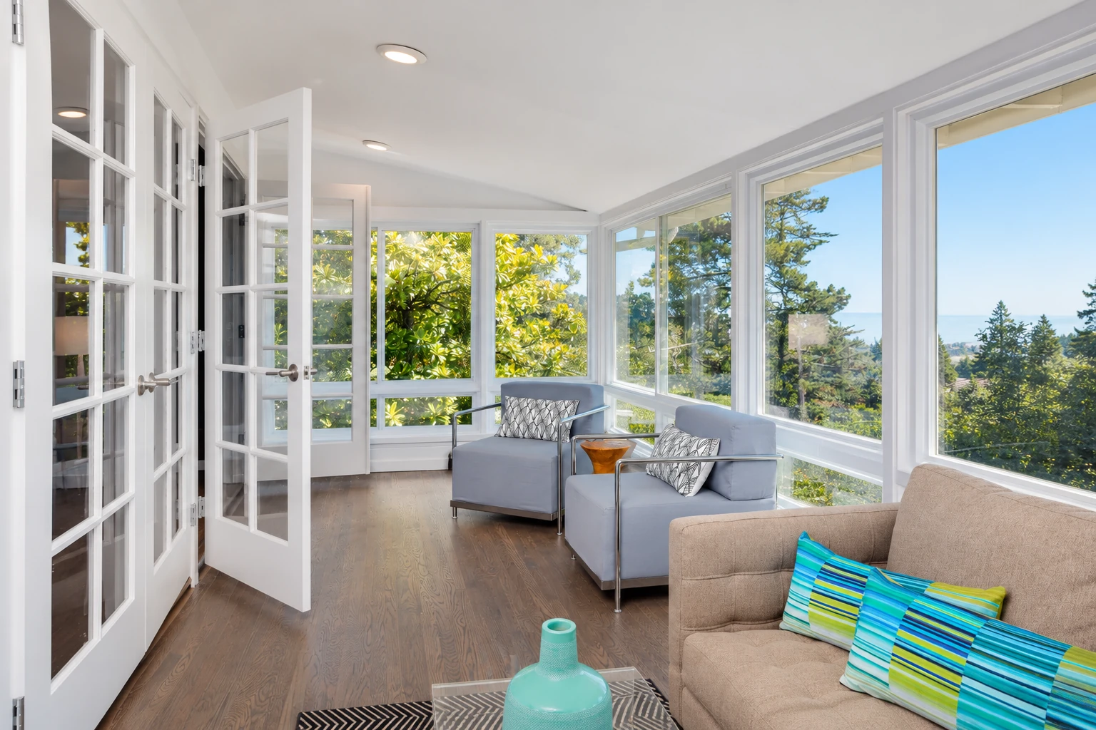 A sunroom with white-framed windows, open French doors, pale blue armchairs, and views of trees and water.