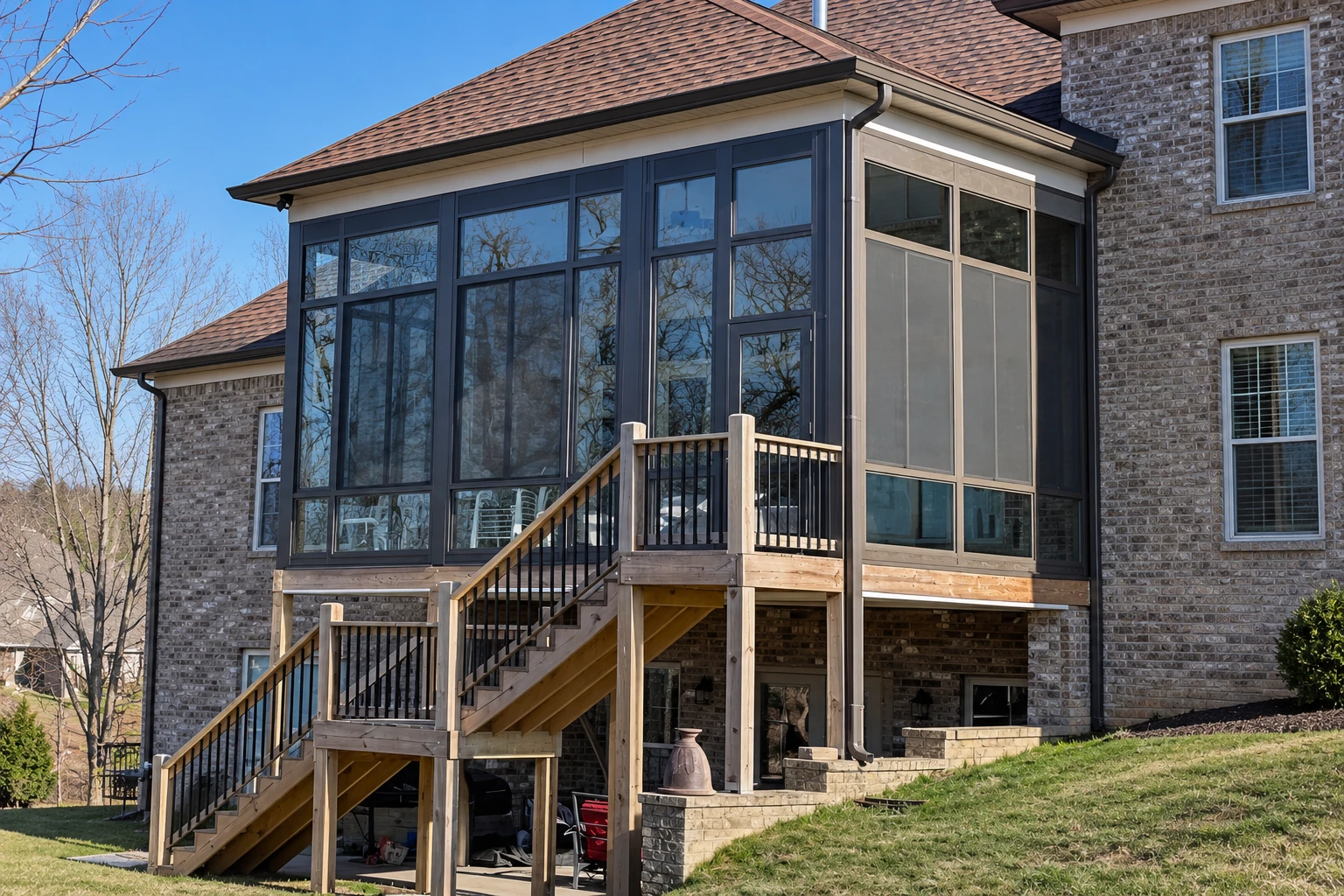Raised sunroom addition with dark-framed glass walls, brick exterior, and a wooden staircase and deck.