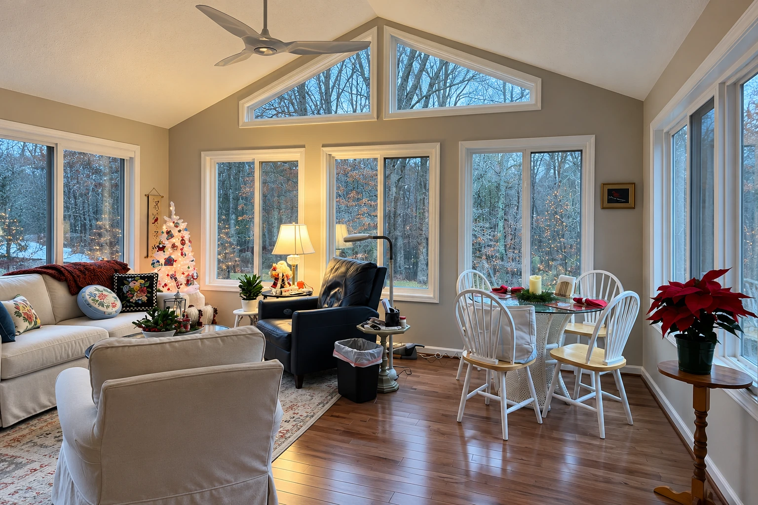 Sunroom interior with tall windows, a ceiling fan, seating area, and dining table overlooking a snowy wooded yard.