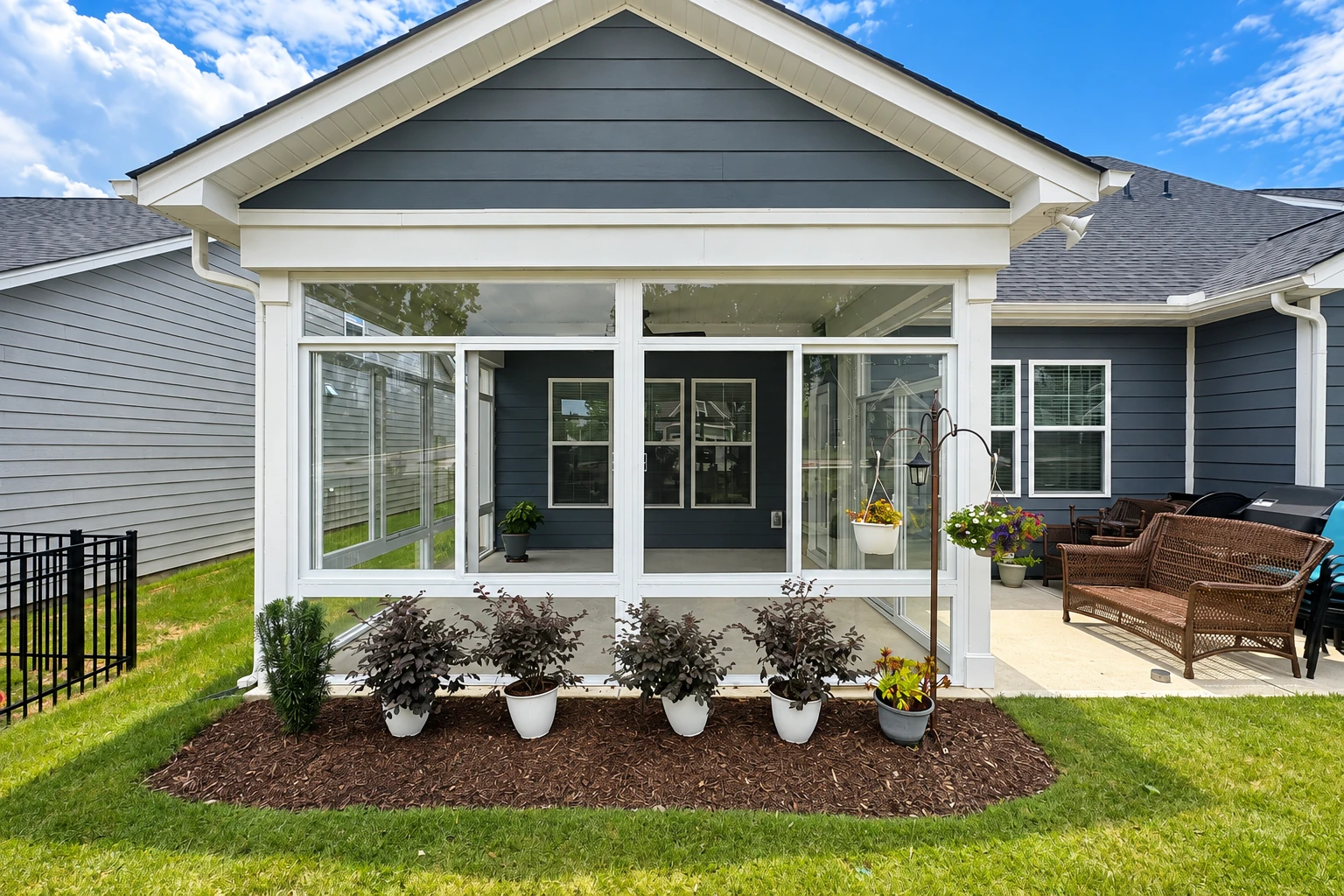 A white-framed three-season sunroom with sliding glass panels sits beside a dark blue house, with potted plants and wicker patio seating nearby.